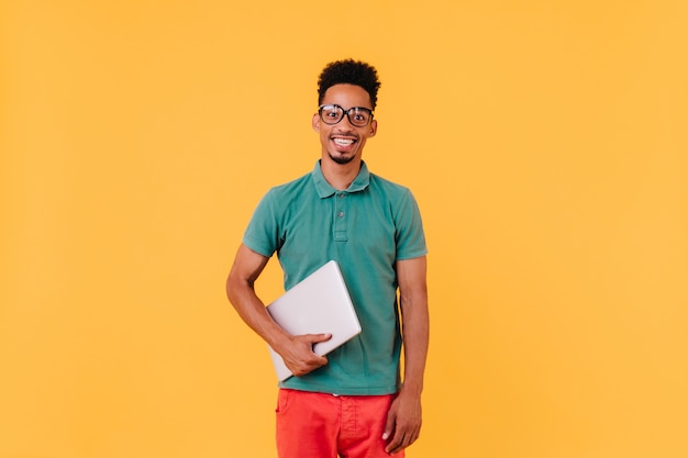 Alegre estudante internacional em t-shirt verde sorrindo. Retrato de freelancer masculino animado com o laptop.