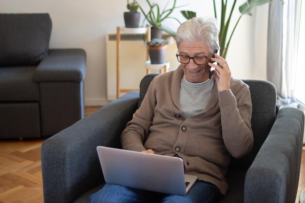 Alegre freelancer idoso trabalhando remotamente. Sorrindo homem grisalho no casaco de lã usando laptop ao ligar por telefone. Conceito de negócios on-line