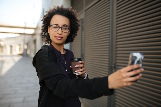 Alegre garota afro-americana tomando selfie e bebendo café na rua, usando óculos