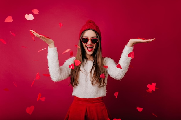 Alegre garota de cabelos compridos com chapéu de malha, se divertindo no dia dos namorados. linda morena de suéter branco em pé sob confetes de coração.