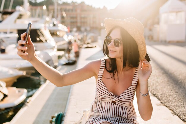 Alegre garota de cabelos escuros em óculos de sol, fazendo selfie enquanto descansava no porto fluvial, aproveitando o sol. Retrato ao ar livre de mulher jovem sorridente com chapéu e vestido tirando foto de si mesma perto de iates.