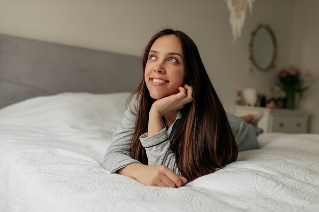 Alegre garota feliz relaxando na cama branca pela manhã. Jovem alegre com longos cabelos castanhos, sonhando e sorrindo.