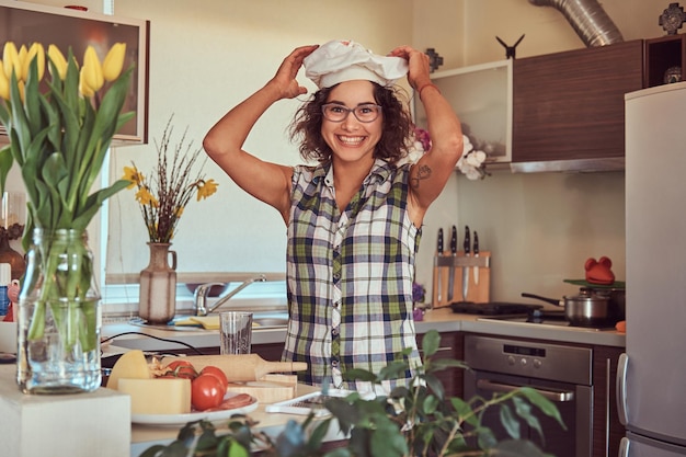 Alegre garota hispânica encaracolada posa na boina de um cozinheiro enquanto cozinha em sua cozinha.