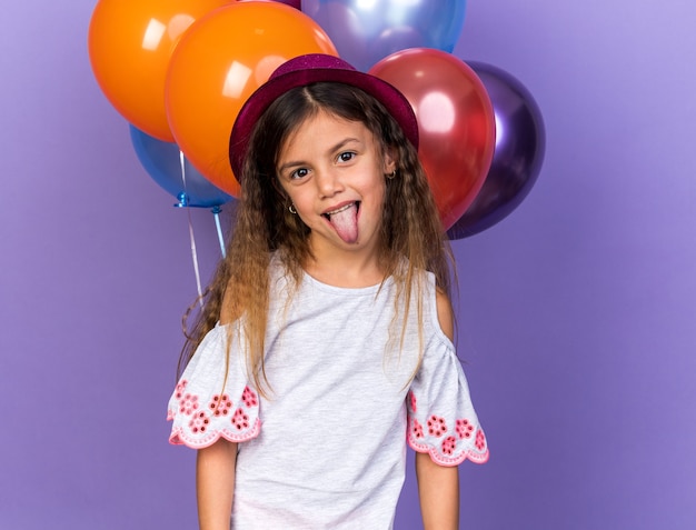Alegre garotinha caucasiana com chapéu de festa violeta levanta a língua em pé na frente de balões de hélio isolados na parede roxa com espaço de cópia