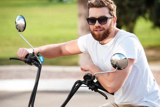 Alegre homem barbudo em óculos de sol, sentado na moto moderna ao ar livre e olhando para a câmera