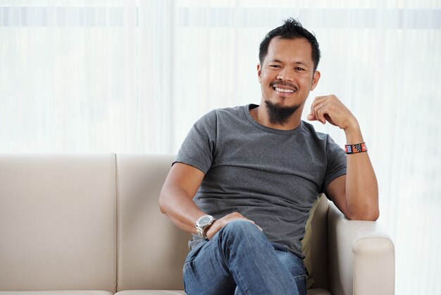 Alegre homem filipino sentado no sofá sorrindo contente para a câmera