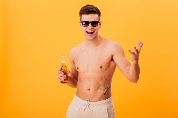 Alegre homem nu de Bermuda e óculos escuros, segurando a garrafa de cerveja