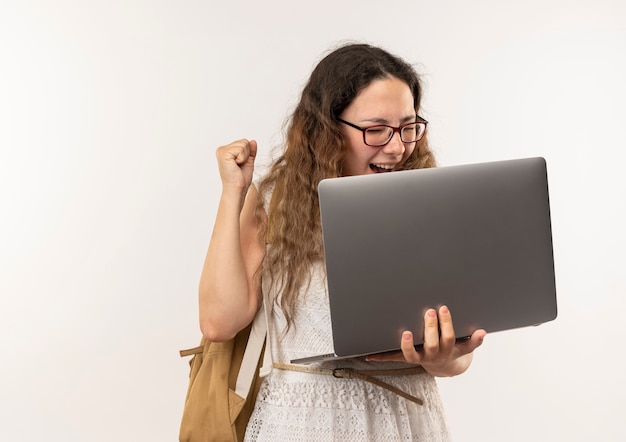 Alegre jovem bonita colegial usando óculos e bolsa traseira segurando laptop levantando o punho isolado