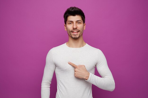 Alegre jovem bonito olhando para a câmera apontando para si mesmo no fundo roxo