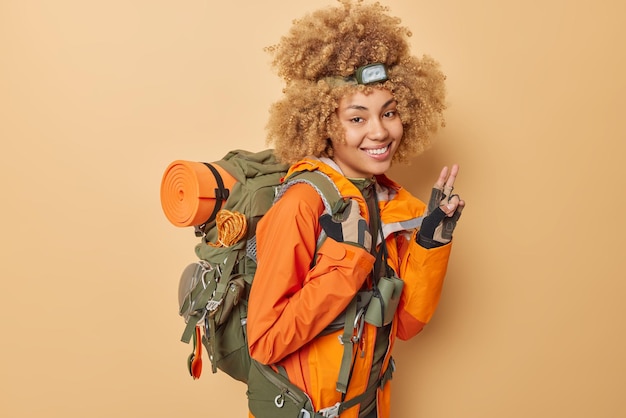 Alegre jovem campista feminina vestida com jaqueta laranja faz gesto de paz desfruta de estilo de vida ativo carrega mochila de bom humor isolado sobre fundo bege caminhadas e conceito de viagem