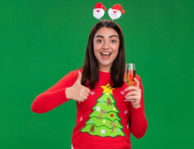 Alegre jovem caucasiana com fita de papai noel, polegares para cima e segurando uma taça de champanhe isolada em um fundo verde com espaço de cópia