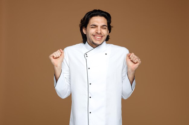 alegre jovem chef masculino vestindo uniforme, mostrando o gesto sim com os olhos fechados, isolados no fundo marrom