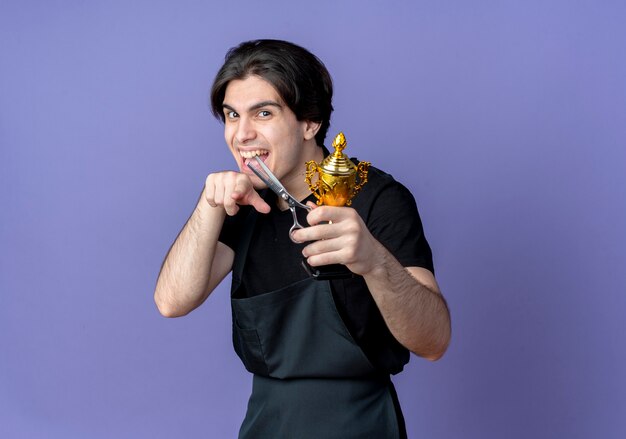 Alegre jovem e bonito barbeiro de uniforme segurando a taça do vencedor com uma tesoura e mostrando um gesto isolado sobre fundo azul