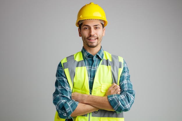 Alegre jovem engenheiro masculino usando capacete de segurança e uniforme olhando para a câmera mantendo os braços cruzados isolados no fundo branco