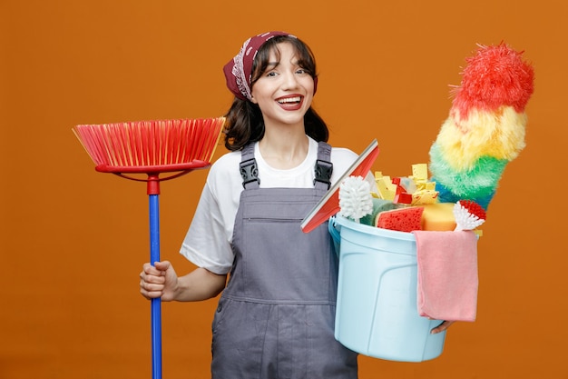 Alegre jovem limpador feminino vestindo uniforme e bandana segurando o esfregão de rodo e balde de ferramentas de limpeza olhando para câmera isolada em fundo laranja