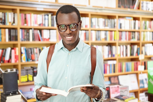 Alegre jovem negro elegante usando óculos em pé na biblioteca com prateleiras de livros