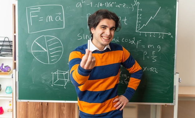 Alegre jovem professor de geometria em frente ao quadro-negro na sala de aula, com as mãos na cintura, olhando para a frente, esticando o giz para frente