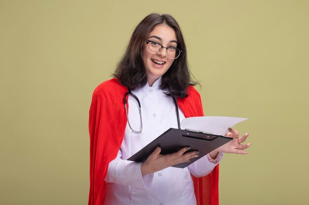 Alegre jovem super-heroína caucasiana com capa vermelha, uniforme de médico e estetoscópio com óculos segurando uma prancheta