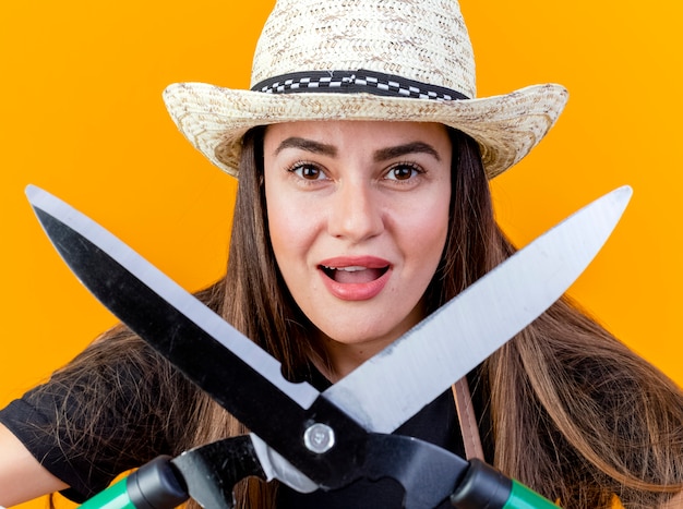 Alegre linda garota jardineira vestindo uniforme e chapéu de jardinagem com luvas segurando uma tesoura para a câmera isolada em fundo laranja