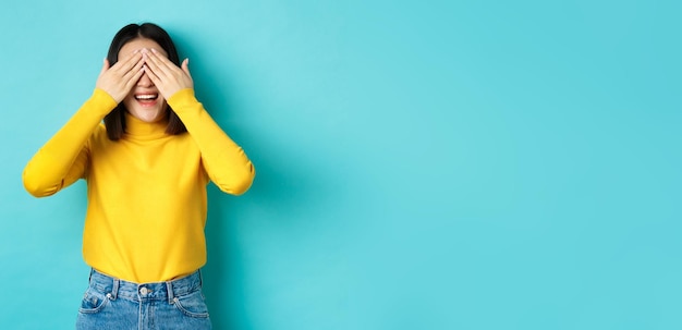 Foto grátis alegre menina asiática de pulôver amarelo esperando surpresa brincando de esconde-esconde e sorrindo esperando