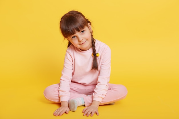 Alegre menina atraente sentada no chão com as pernas cruzadas, tocando o chão com as palmas das mãos, olhando para a câmera, posando isolado sobre fundo amarelo, vestidos de traje rosa pálido.