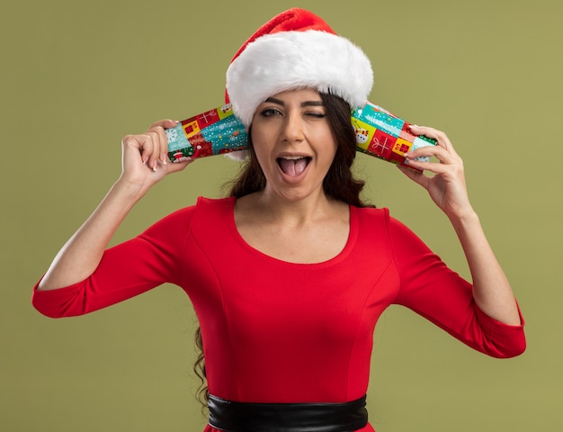 Foto grátis alegre menina bonita com chapéu de papai noel segurando xícaras de café de natal perto de orelhas, ouvindo conversas piscando isoladas na parede verde-oliva