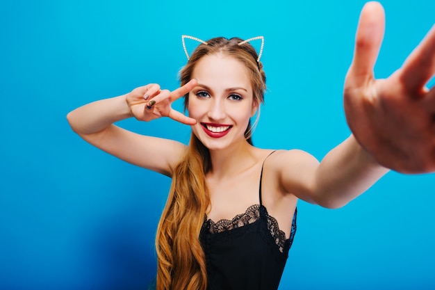 Alegre menina bonita com orelhas de gato em diamantes na cabeça posando, tomando selfie, mostrando paz, curtindo a festa. De vestido preto, tem lindos olhos azuis, cabelos longos e ondulados.