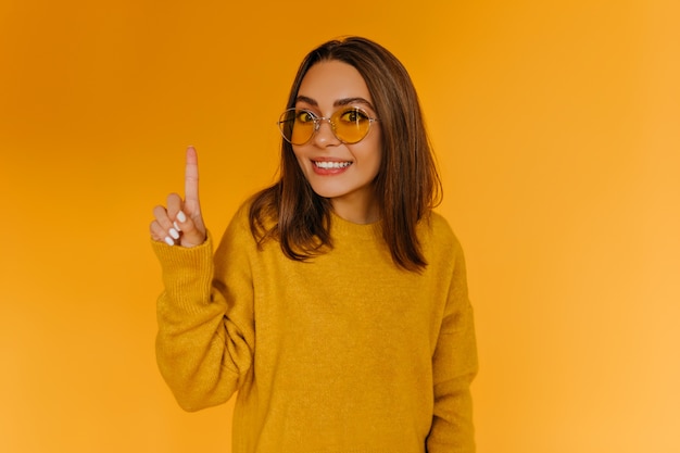 Alegre menina bronzeada em copos posando na parede laranja. jovem atraente numa camisola de malha, brincando.