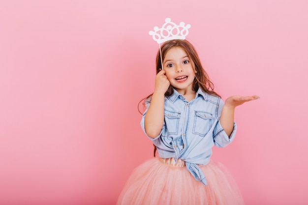 Alegre menina com longos cabelos castanhos em saia de tule, segurando a coroa de princesa na cabeça isolada no fundo rosa. comemorando carnaval brilhante para crianças, expressando positividade da festa de aniversário