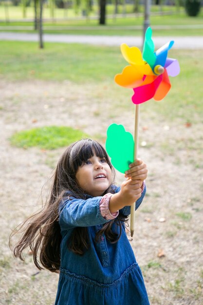 Alegre menina de cabelos negros brincando no parque, segurando e levantando o cata-vento, olhando para o brinquedo de emoção. Tiro vertical. Conceito de atividade infantil ao ar livre
