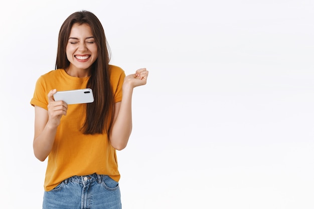 Alegre menina moderna fofa triunfando ao passar de nível no jogo, amigas pontuam no aplicativo móvel, segurando o smartphone horizontalmente, sorrindo com os olhos fechados animado punho cerrado, fundo branco