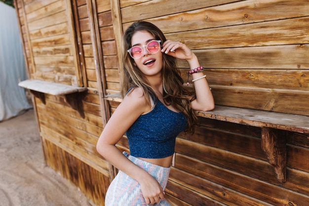 Alegre menina modish em óculos de sol sonhadora posando, encostada na parede do prédio de madeira.
