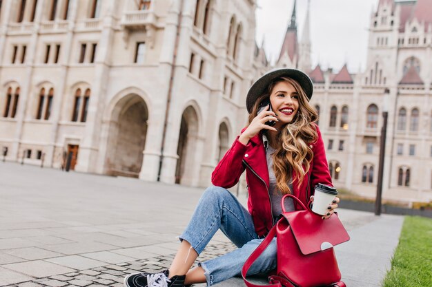 Alegre modelo feminina branca com roupa de rua chamando um amigo enquanto está sentado ao lado do lindo palácio