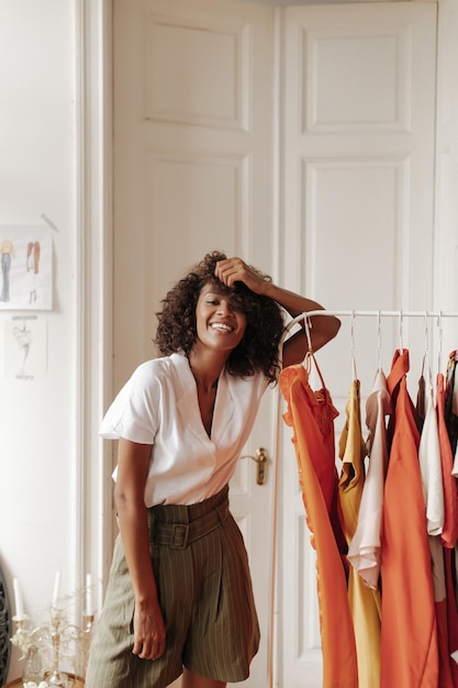 Alegre morena legal morena encaracolada em shorts cáqui e blusa branca sorri toca o cabelo e se inclina no cabide no quarto aconchegante