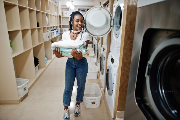 Alegre mulher afro-americana com toalhas nas mãos perto da máquina de lavar roupa na lavanderia self-service