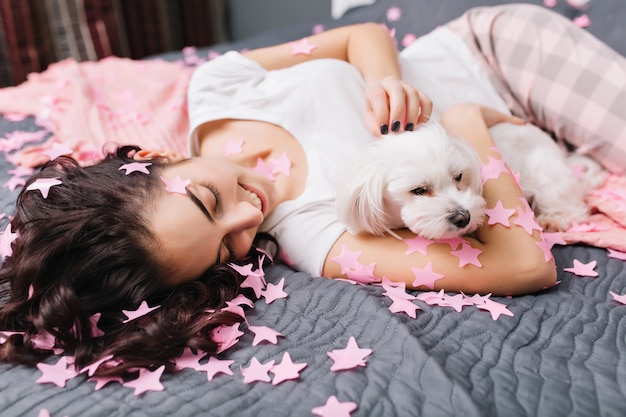 Alegre mulher bonita jovem com cabelo encaracolado morena de pijama, relaxando na cama com o cachorrinho em enfeites rosa. Linda modelo se divertindo em casa com animais domésticos, expressando felicidade