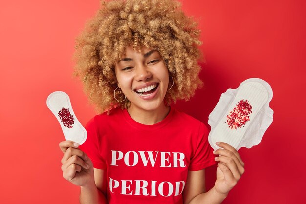 Foto grátis alegre mulher com cabelo espesso encaracolado segura juntas orgânicas feitas de algodão bio ri positivamente vestida com camiseta casual com inscrição isolada sobre fundo vermelho vívido conceito de higiene