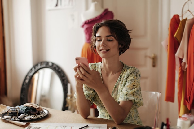 Alegre mulher de cabelos curtos morena bronzeada em vestido floral com decote em v, conversando no telefone, senta-se na mesa e posa no escritório de designer de moda aconchegante