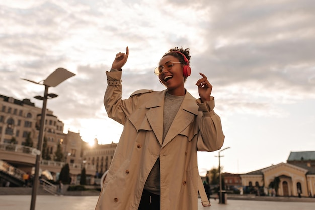 Foto grátis alegre mulher de pele escura em trench coat bege de grandes dimensões e sorrisos de óculos ouve música e dança ao ar livre