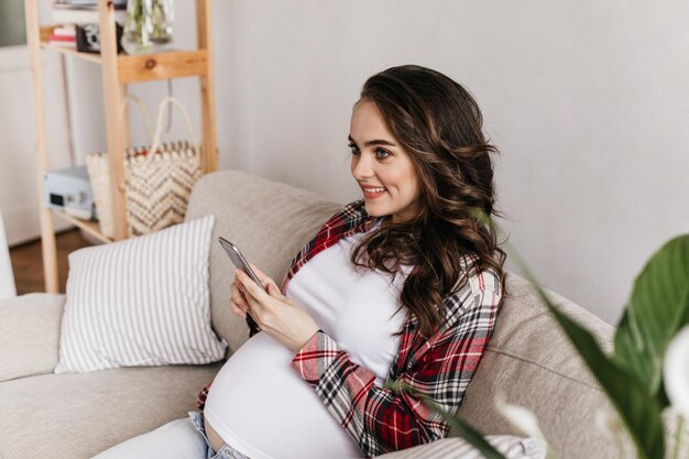 Alegre mulher grávida morena de ótimo humor sorri e descansa no sofá macio Jovem mãe segura telefone e posa na aconchegante sala de estar