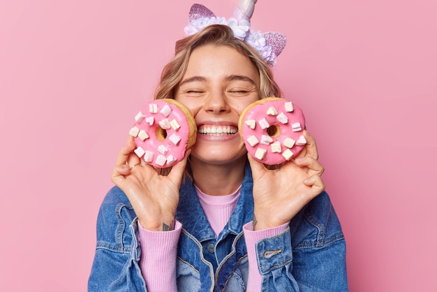 Alegre mulher jovem de cabelos louros mantém os olhos fechados sorrisos dentuça segura dois donuts com marshmallow feliz em comer a sobremesa favorita vestida com jaqueta jeans isolada sobre fundo rosa