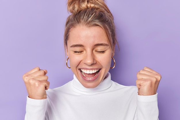 Alegre mulher otimista com coque de cabelo mantém os olhos fechados sorri amplamente cerra os punhos exclama de alegria celebra boas notícias usa jumper branco isolado sobre fundo roxo ganha e se alegra