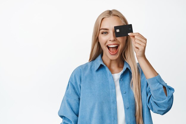 Alegre mulher sorridente segurando o cartão de crédito contra os olhos gritando de pé feliz sobre fundo branco