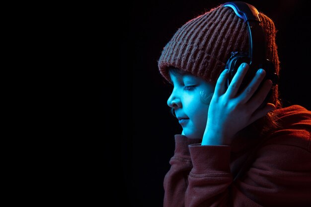 Alegre ouvindo música em fones de ouvido com os olhos fechados. Retrato do menino caucasiano em fundo escuro em luz de néon. Conceito de emoções humanas, expressão facial, vendas, anúncio, tecnologia moderna, gadgets.