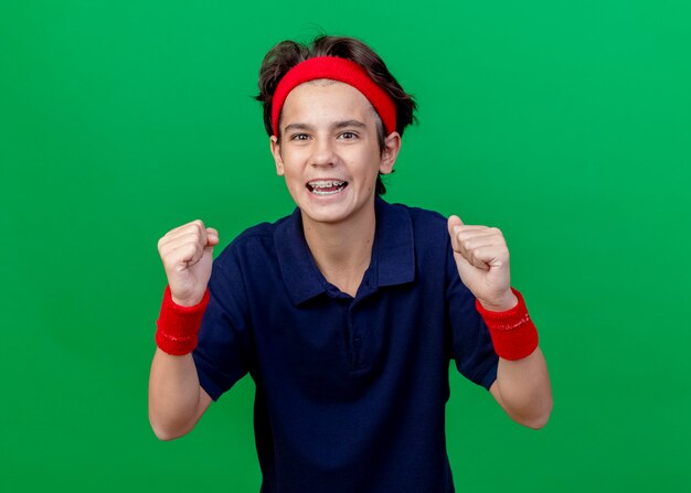 Alegre rapaz bonito desportivo usando bandana e pulseiras com aparelho dentário, olhando para a câmera, fazendo gesto de sim isolado no fundo verde
