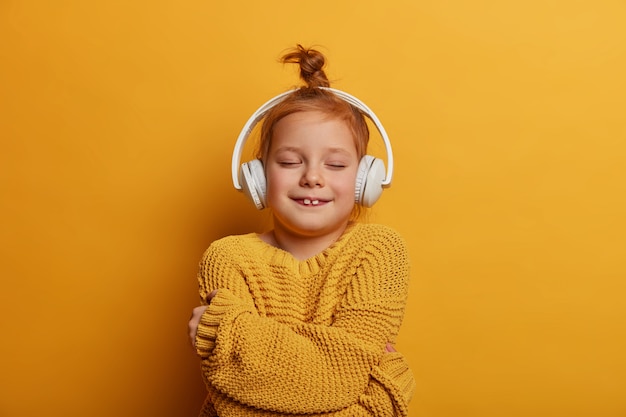 Alegre ruiva com coque de cabelo, se abraça, sente-se confortável com o novo suéter de tricô, ouve melodia em fones de ouvido estéreo, isolados sobre vibrante parede amarela. crianças, conceito de passatempo