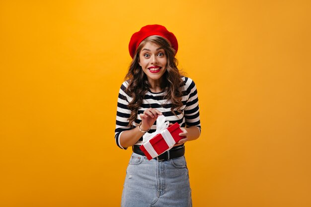 Alegre senhora de boina vermelha segura caixa de presente em fundo laranja. Linda mulher com cabelo encaracolado de suéter listrado e saia jeans, olhando para a câmera.