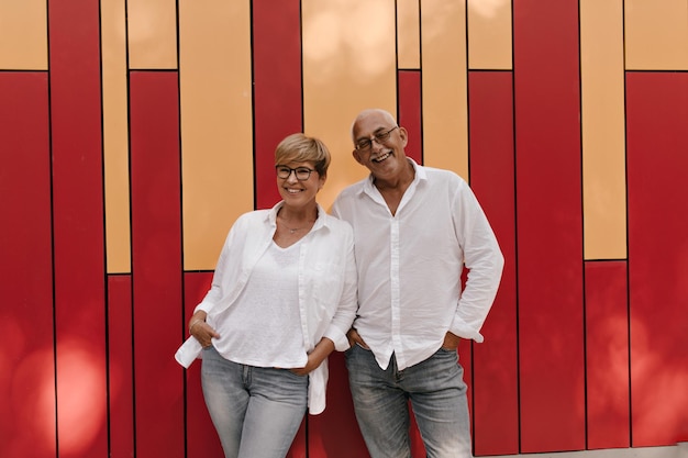 Alegre senhora de cabelo loira de camisa branca e jeans sorrindo e posando com homem de óculos com roupas leves em pano de fundo vermelho e laranja