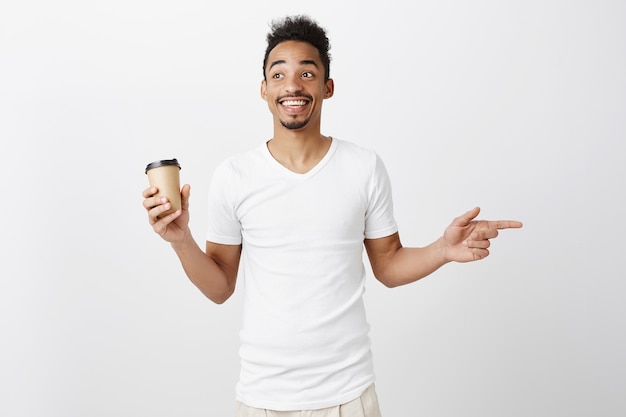 Alegre sorridente cara afro-americana com camiseta branca apontando para a direita, segurando a xícara com café