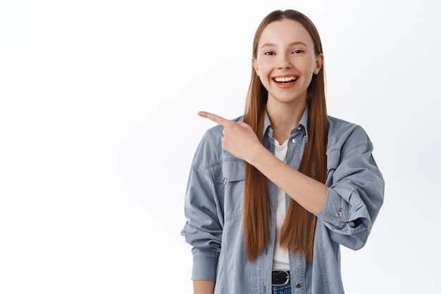 Alegre universitária com cabelos compridos apontando para o lado sorrindo e mostrando o logotipo dessa forma gesto recomendando clicar no banner rindo feliz em pé sobre fundo branco